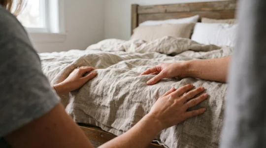 Une personne prépare un lit en installant du linge en lin sur un matelas équipé d'un sur-matelas à mémoire de forme visible, montrant les différentes couches du système de couchage dans une chambre résidentielle
